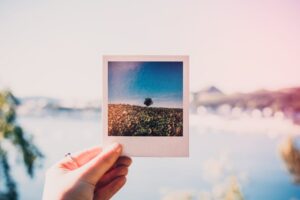 A hand holds a Polaroid picture against a blurred, sunlit summer landscape, creating a beautiful contrast.